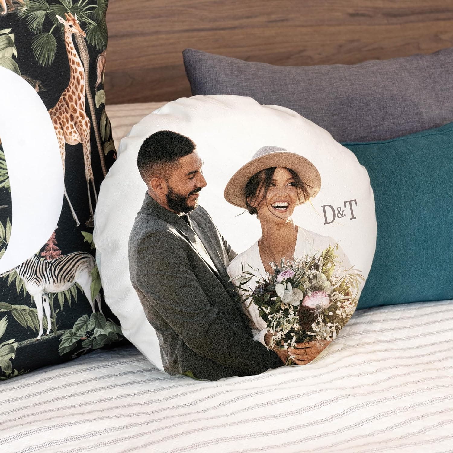 Cojín redondo con la foto de una pareja de novios y las iniciales «D&T», apoyado entre otros cojines en una cama