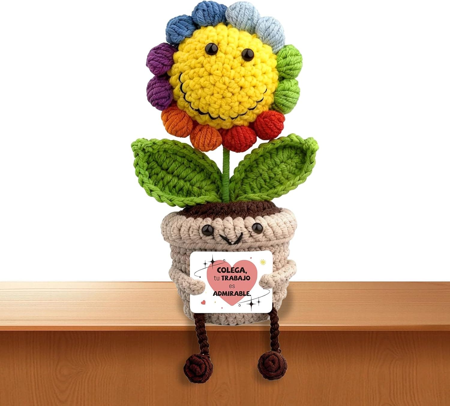 Girasol de ganchillo con maceta que tiene ojos y sonrisa, sentado en un borde con las piernas colgando y sosteniendo una tarjeta con el mensaje "colega, tu trabajo es admirable"