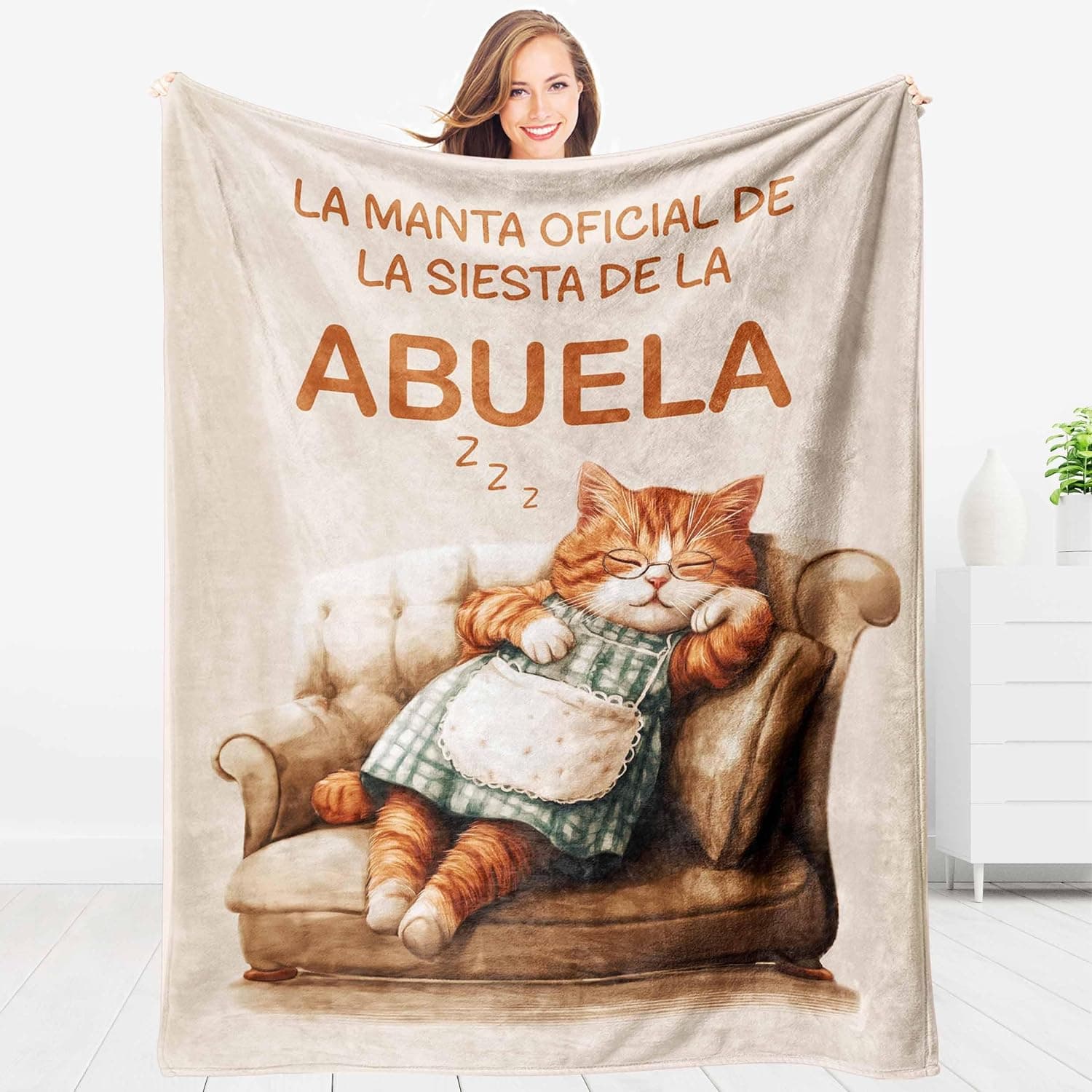 Manta beige con un gato naranja vestido de abuela dormido en un sillón y el texto "la manta oficial de la siesta de la abuela", sostenida por una mujer