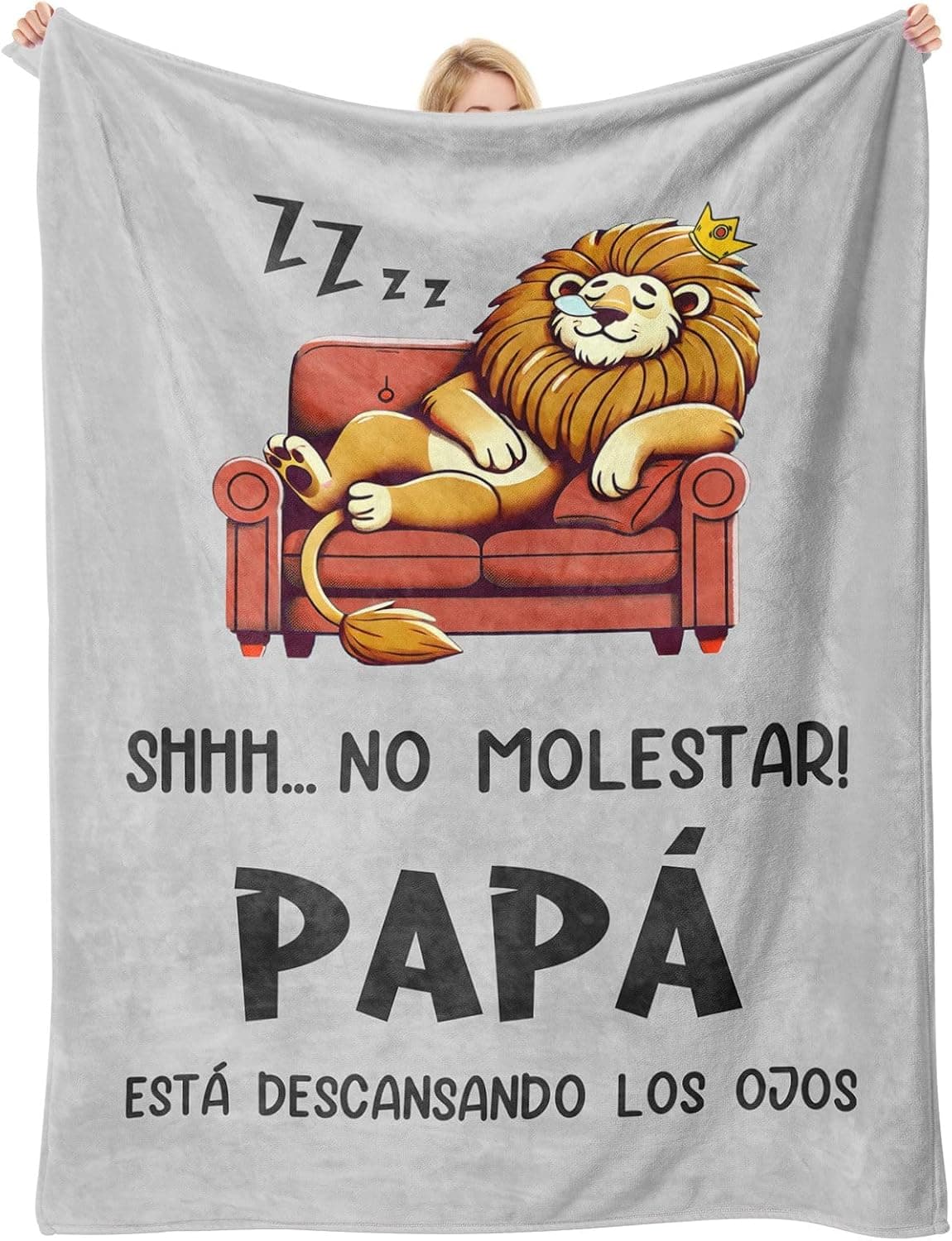 Manta gris con un león dormido en un sofá y el texto "shhh, no molestar, papá está descansando los ojos", sostenida por una mujer