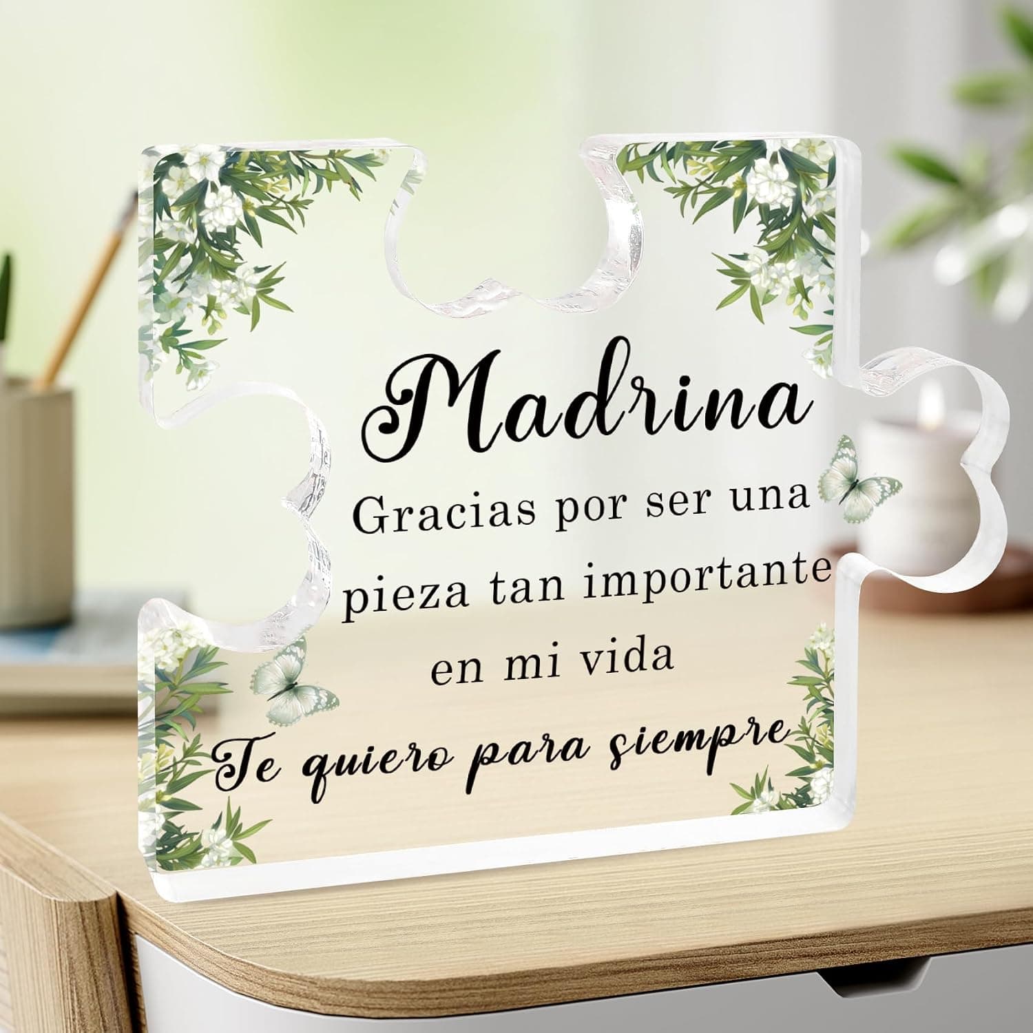 Pieza de puzle de metacrilato transparente de pie sobre una mesa de madera, impresa con «Madrina, gracias por ser una pieza tan importante en mi vida, te quiero para siempre», con ramas de flores blancas en las esquinas y dos mariposas verdes