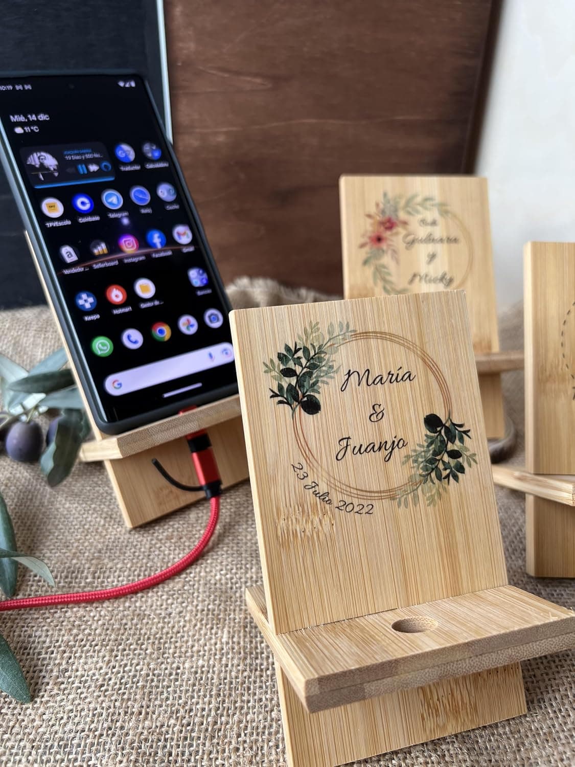 Cuatro soportes de madera para móvil grabados con nombres distintos y la fecha del evento, sobre una mesa con ramas de eucalipto