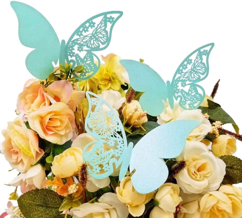 Tarjeta con forma de mariposa turquesa encajada en el borde de una copa de vino vacía, con un nombre escrito a mano en el ala, sobre una mesa de madera con rosas