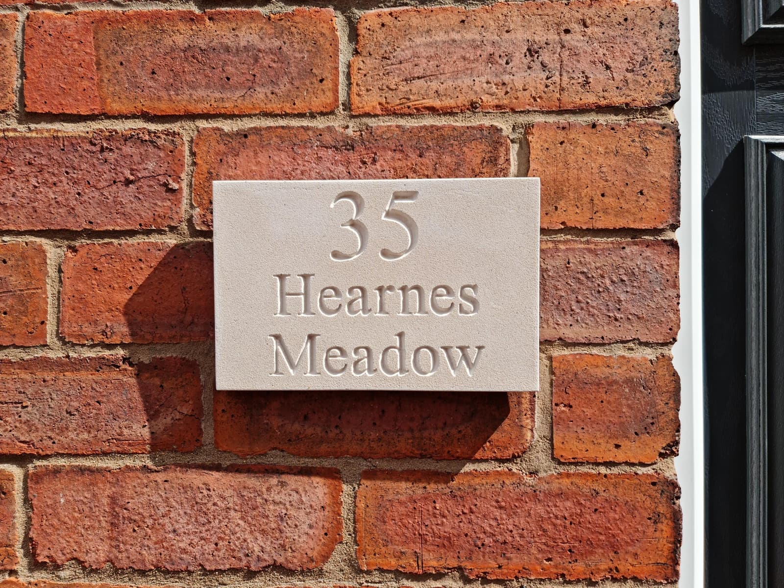 Natural stone house sign with deep V-cut engraving showing '35 Hearnes Meadow' mounted on a red brick wall. The off-white Jesmonite plaque features a fine-grained, matte texture mimicking limestone