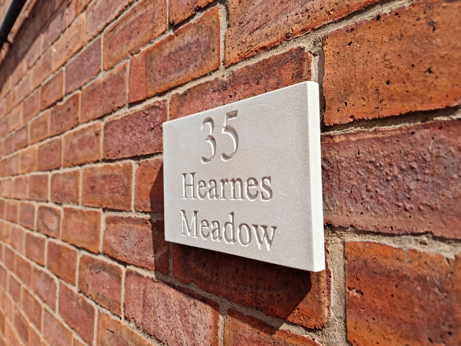 Natural stone house sign with deep V-cut engraving showing '35 Hearnes Meadow' mounted on a red brick wall. The off-white Jesmonite plaque features a fine-grained, matte texture mimicking limestone