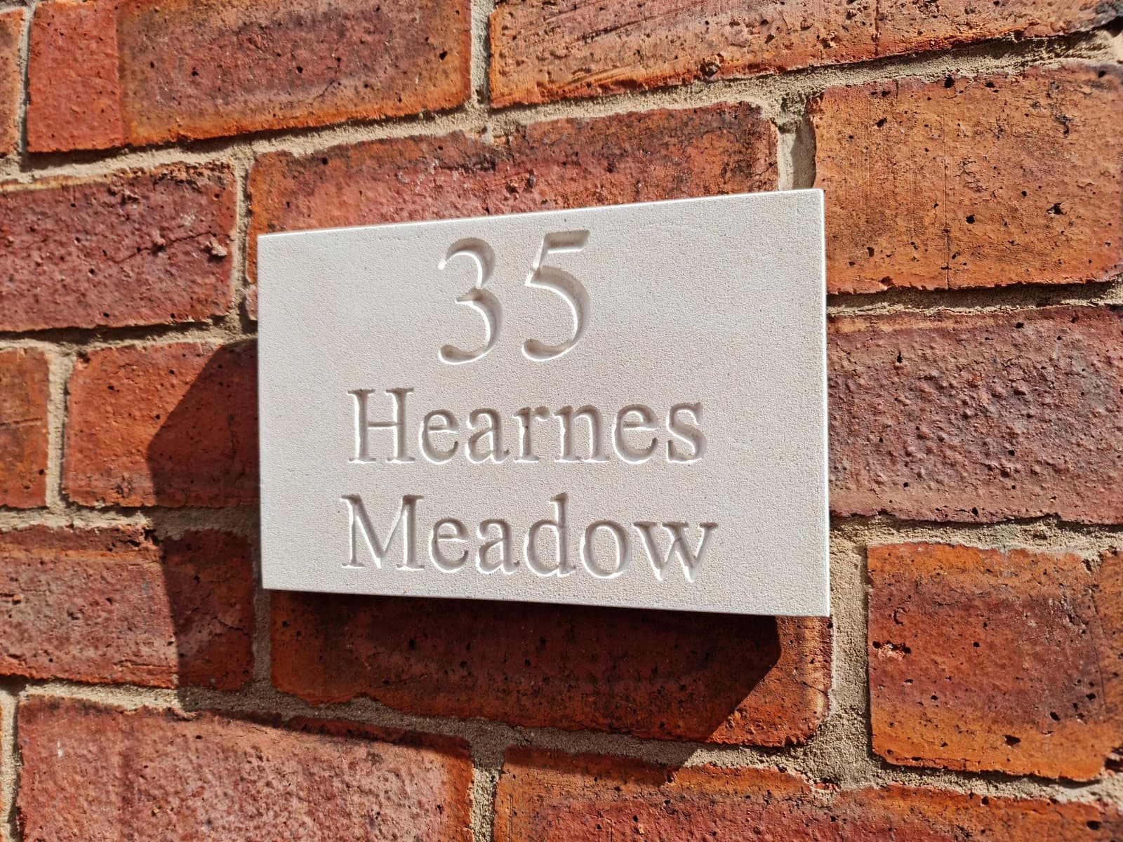 Natural stone house sign with deep V-cut engraving showing '35 Hearnes Meadow' mounted on a red brick wall. The off-white Jesmonite plaque features a fine-grained, matte texture mimicking limestone