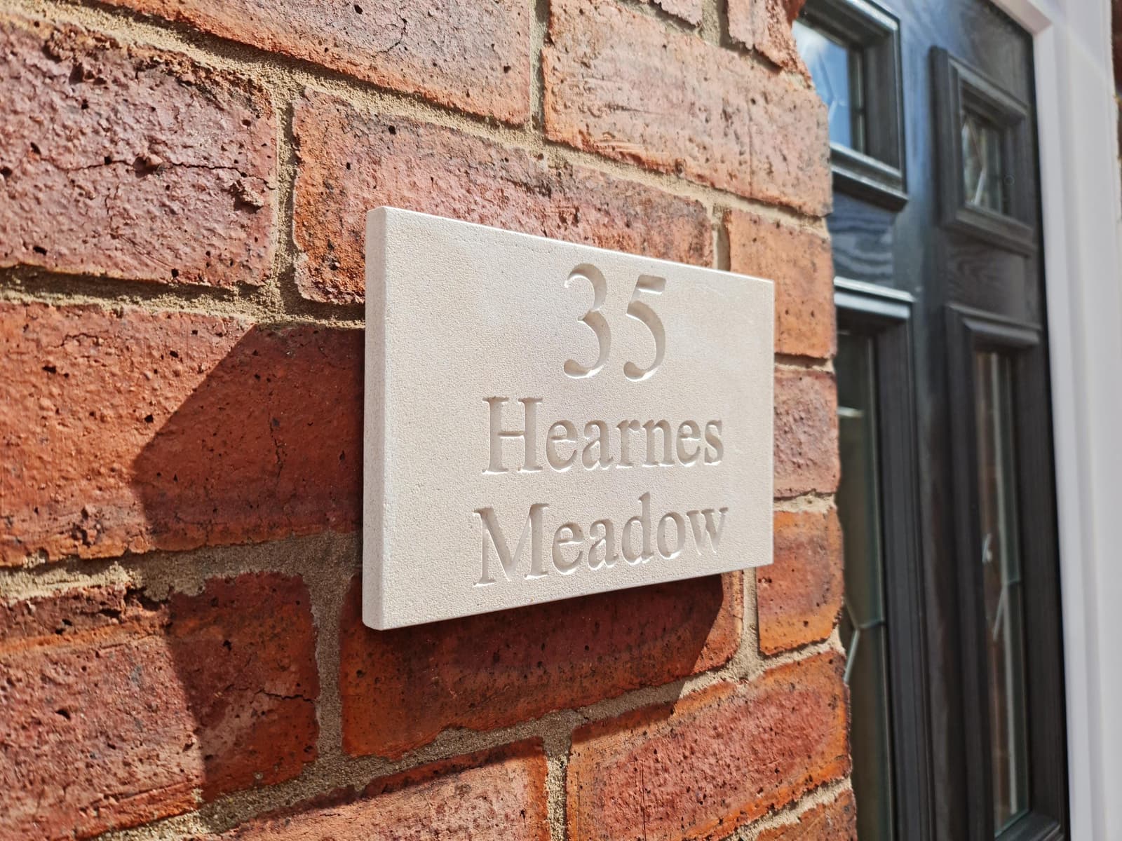 Natural stone house sign with deep V-cut engraving showing '35 Hearnes Meadow' mounted on a red brick wall. The off-white Jesmonite plaque features a fine-grained, matte texture mimicking limestone