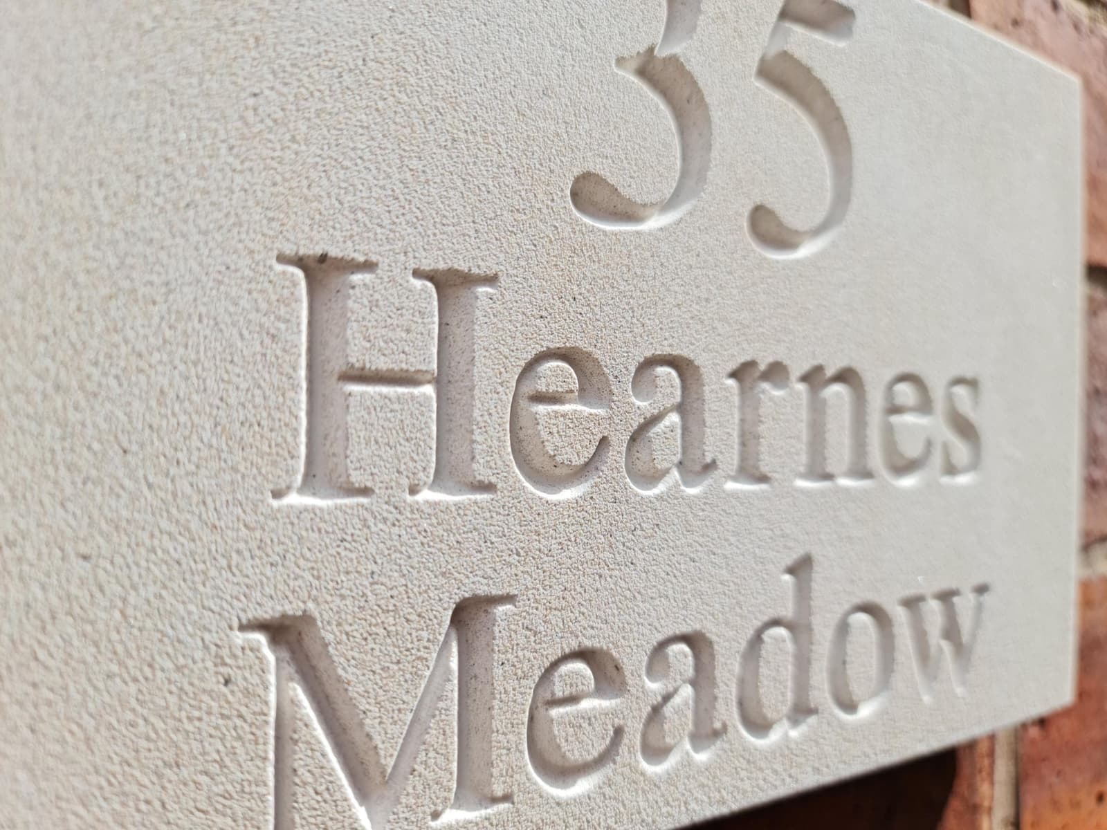 Natural stone house sign with deep V-cut engraving showing '35 Hearnes Meadow' mounted on a red brick wall. The off-white Jesmonite plaque features a fine-grained, matte texture mimicking limestone