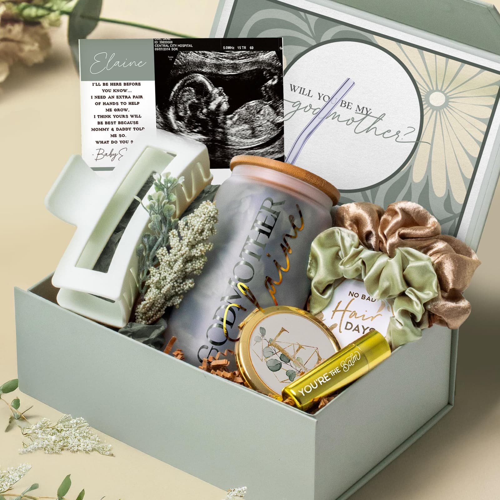 May include: A gift box with baby-themed items. Includes a glass jar with "Godmother" text, a hair clip, scrunchies, a compact mirror, a small tube, a card with text, and a sonogram image. The box is light green.