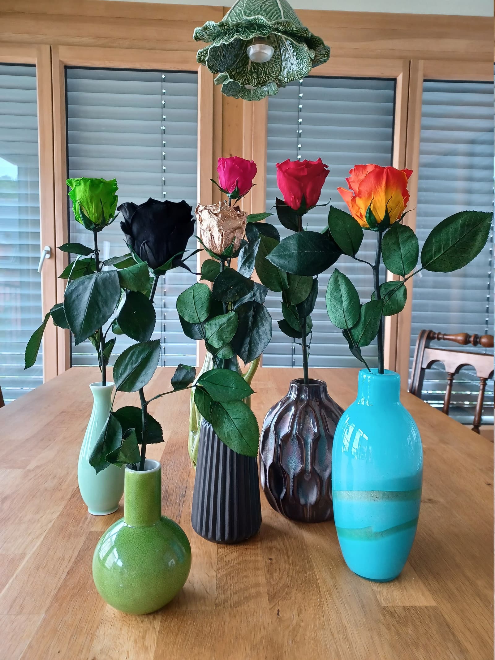 Sechs farbige Infinity-Rosen (Grün, Schwarz, Gold, Pink, Rot, Orange) in verschiedenen Vasen auf Holztisch – langlebige Deko.