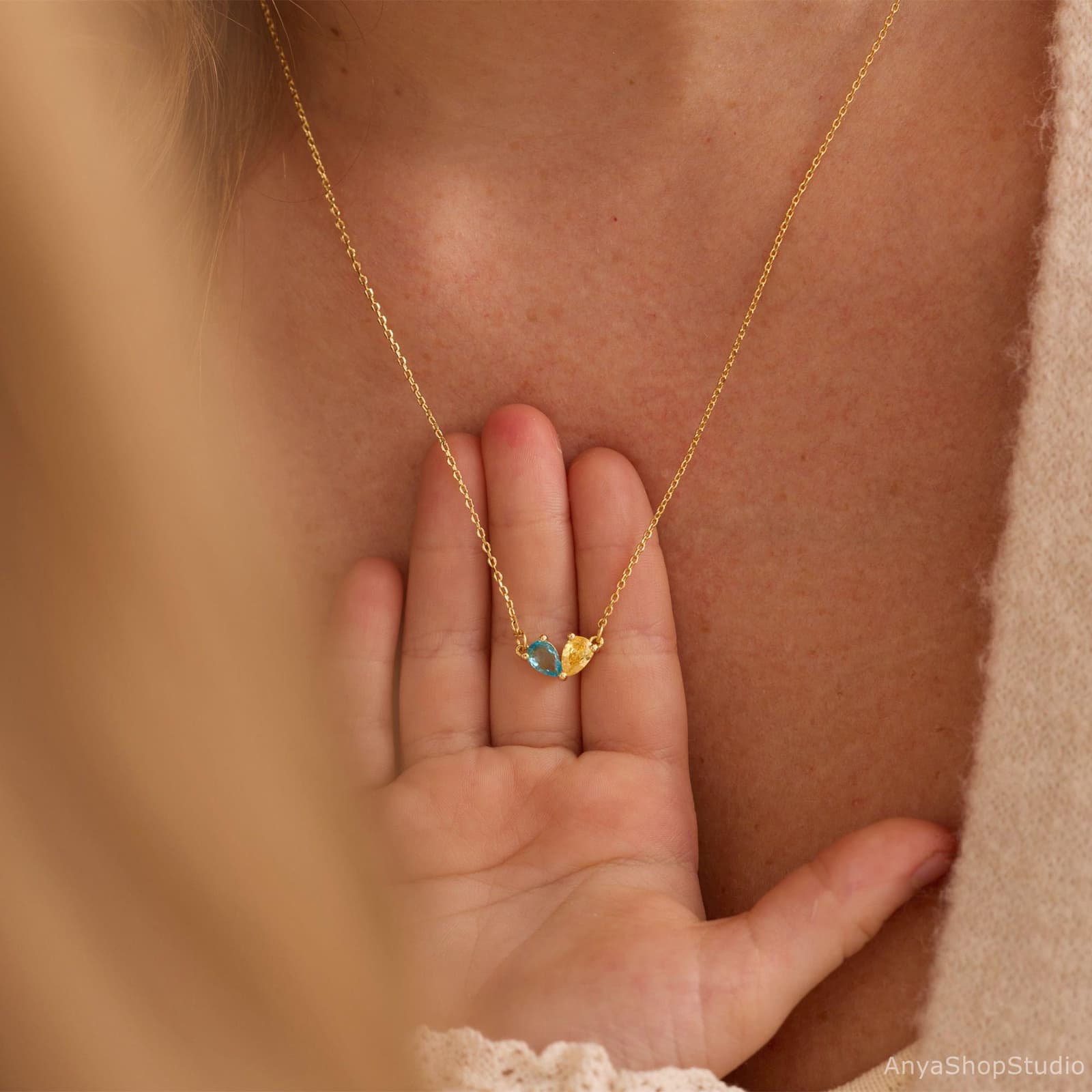 May include: A gold necklace with a fine chain, showcasing two teardrop-shaped gemstones, one blue and one yellow. The necklace is worn on a person's chest, with the pendant resting on their hand.