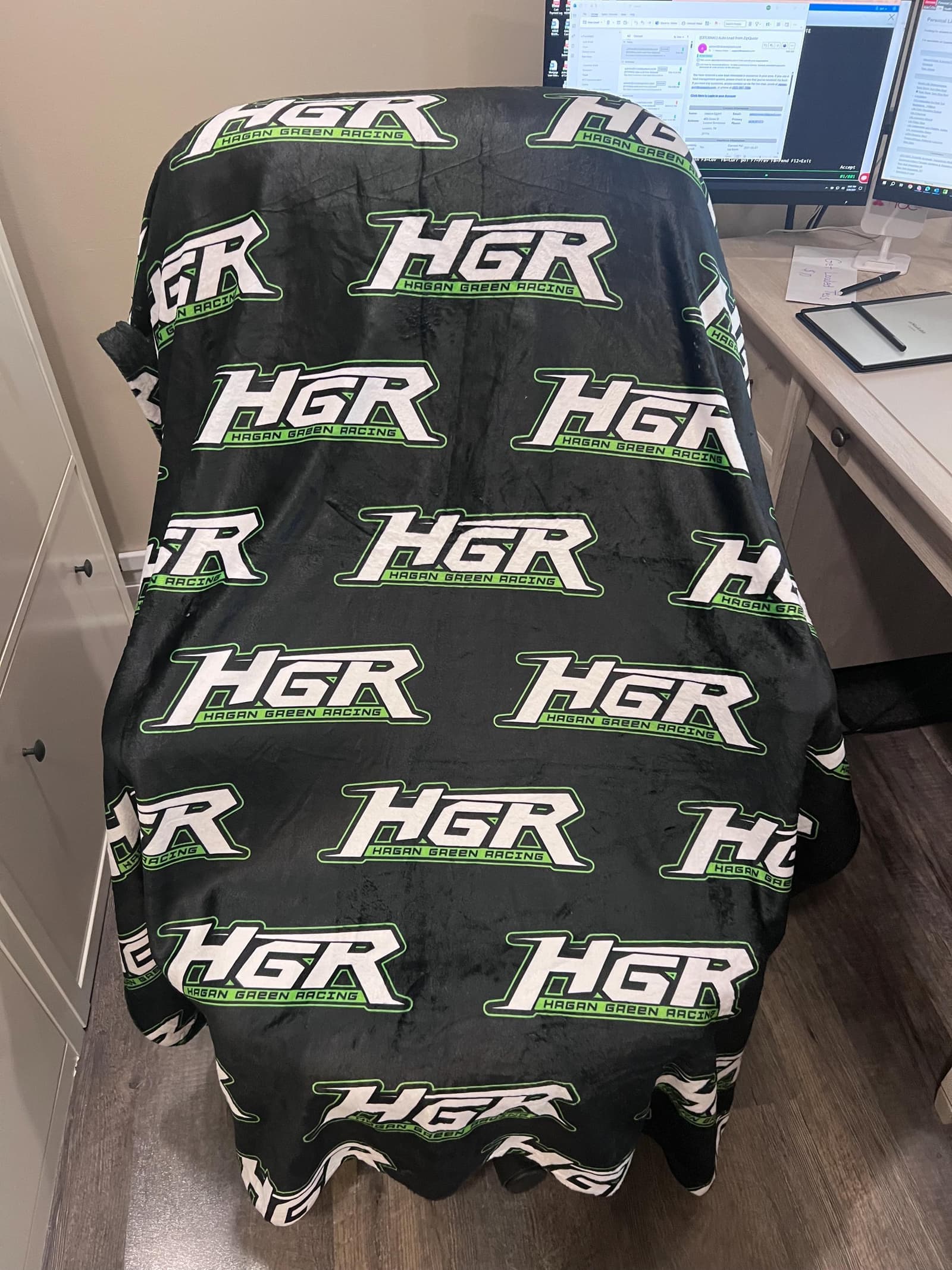 a black blanket with the repeated text "HGR" in white and green. The blanket is draped over a chair, and there is a computer monitor and a keyboard visible in the background.