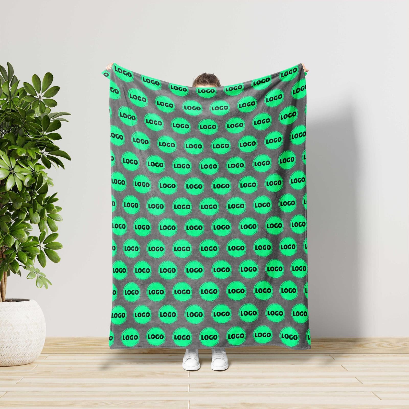 A person is standing in front of a wall, holding a large blanket with a repeating pattern of green circles and the word "LOGO" printed on it.