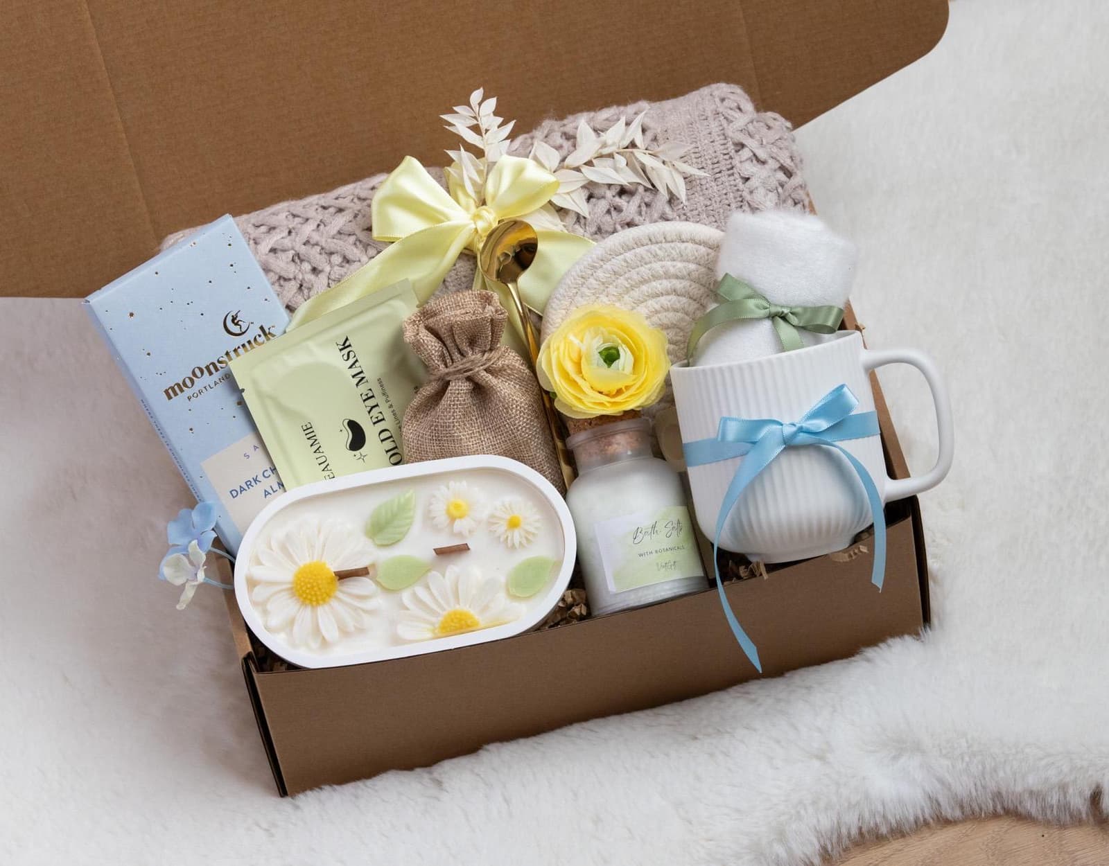 May include: A gift box filled with self-care items. Includes a white mug with a blue ribbon, a candle with daisy accents, bath salts, eye masks, and a yellow flower. The box is lined with a soft, neutral-colored blanket.