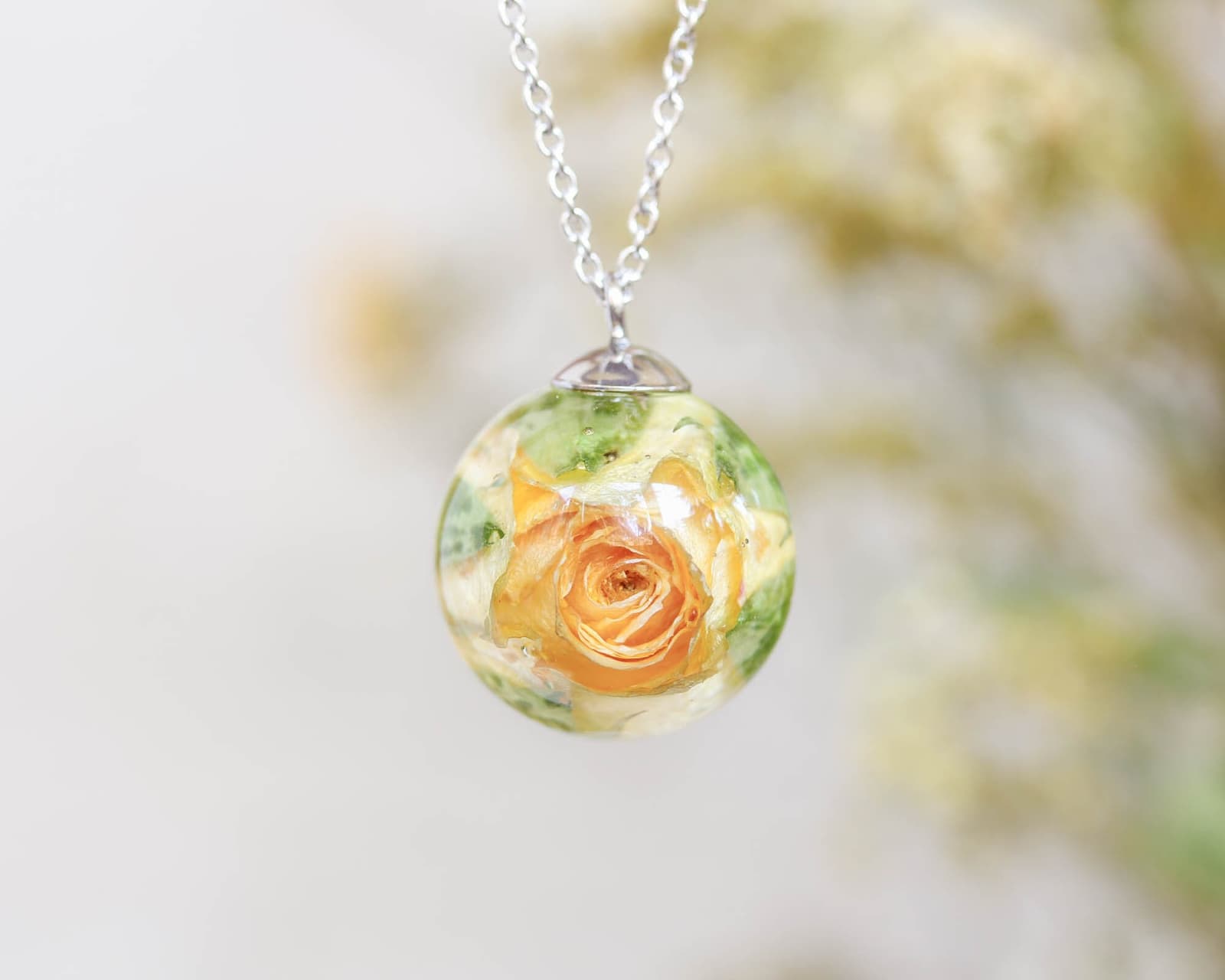Close-up of a handmade resin pendant with a preserved yellow rose and green foliage inside, hanging from a silver chain.
