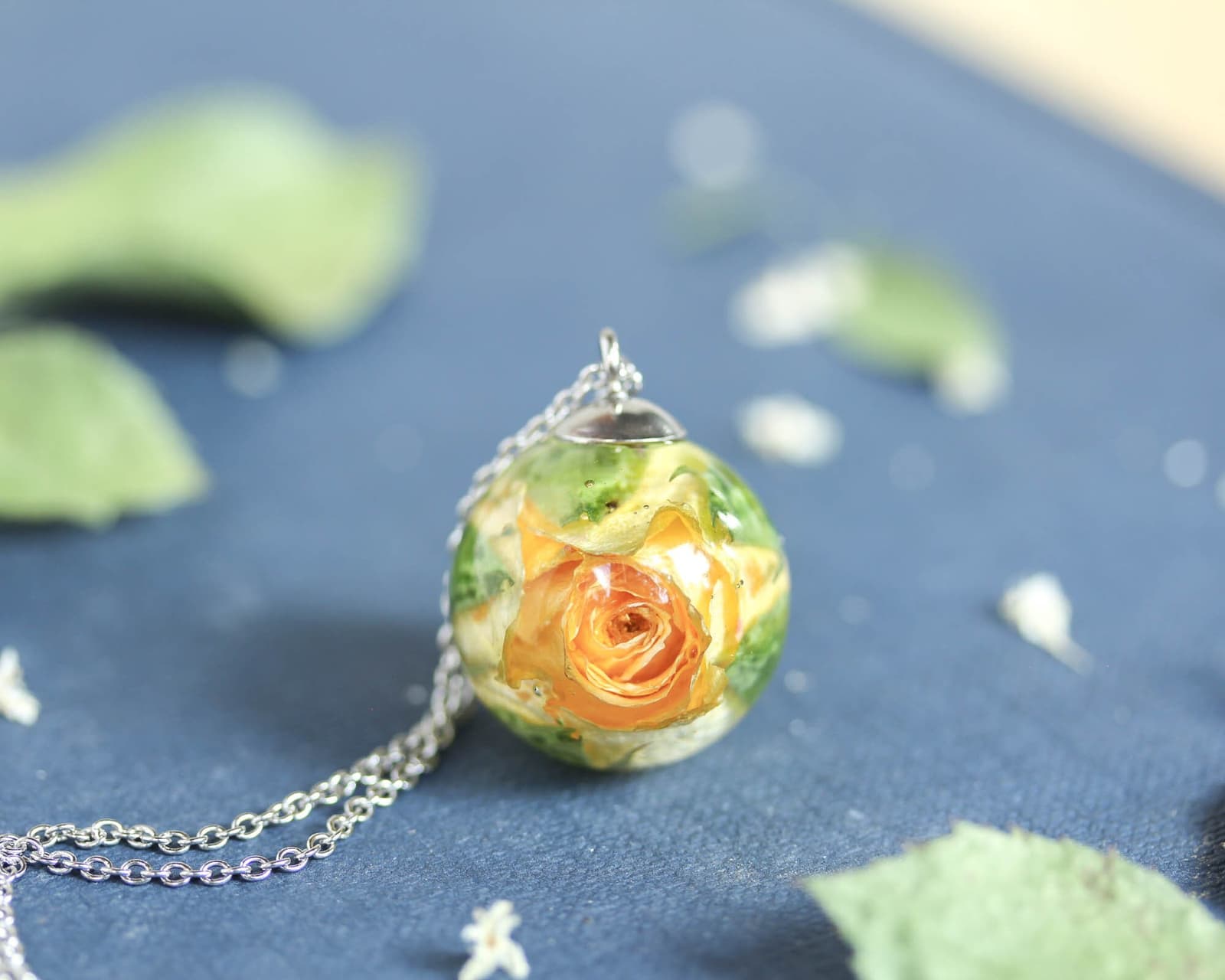Yellow rose pendant resting on a dark surface with a silver chain, surrounded by soft natural elements.