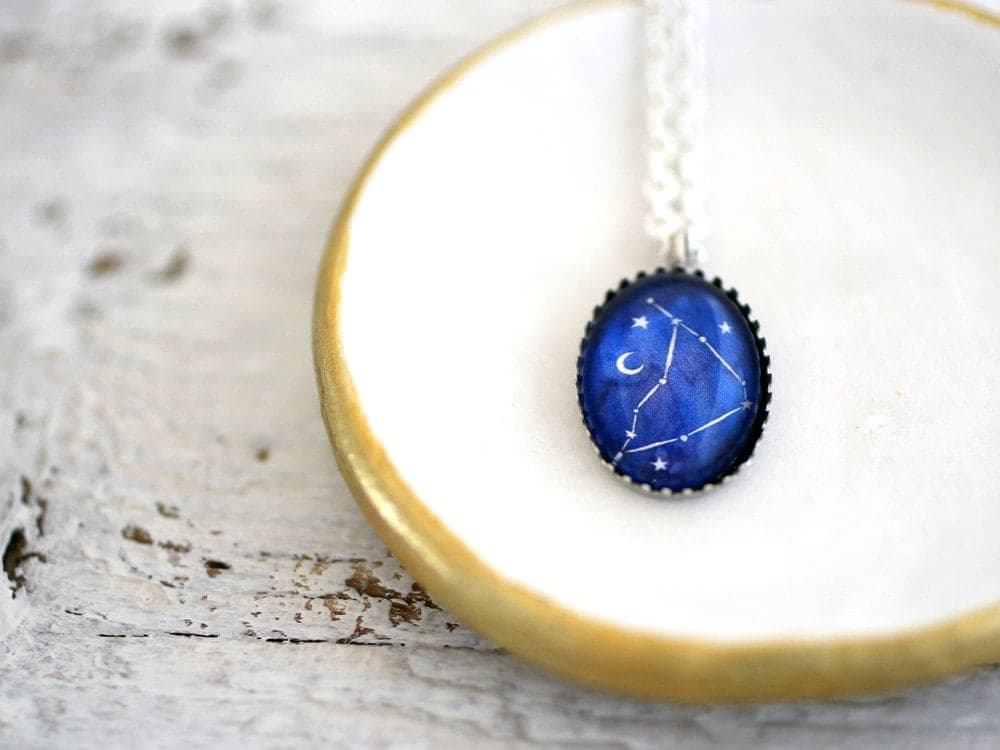 Colgante ovalado con la constelación de Capricornio pintada sobre fondo azul noche con una luna creciente