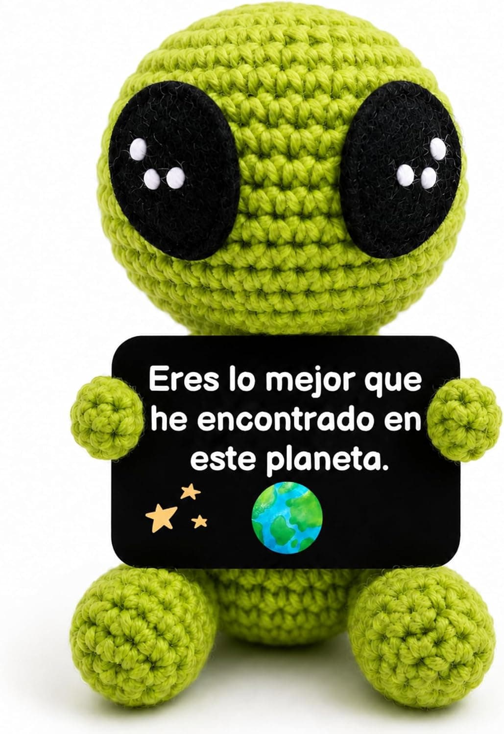 Peluche verde de ganchillo con forma de alienígena y ojos negros grandes, sosteniendo una tarjeta con el mensaje