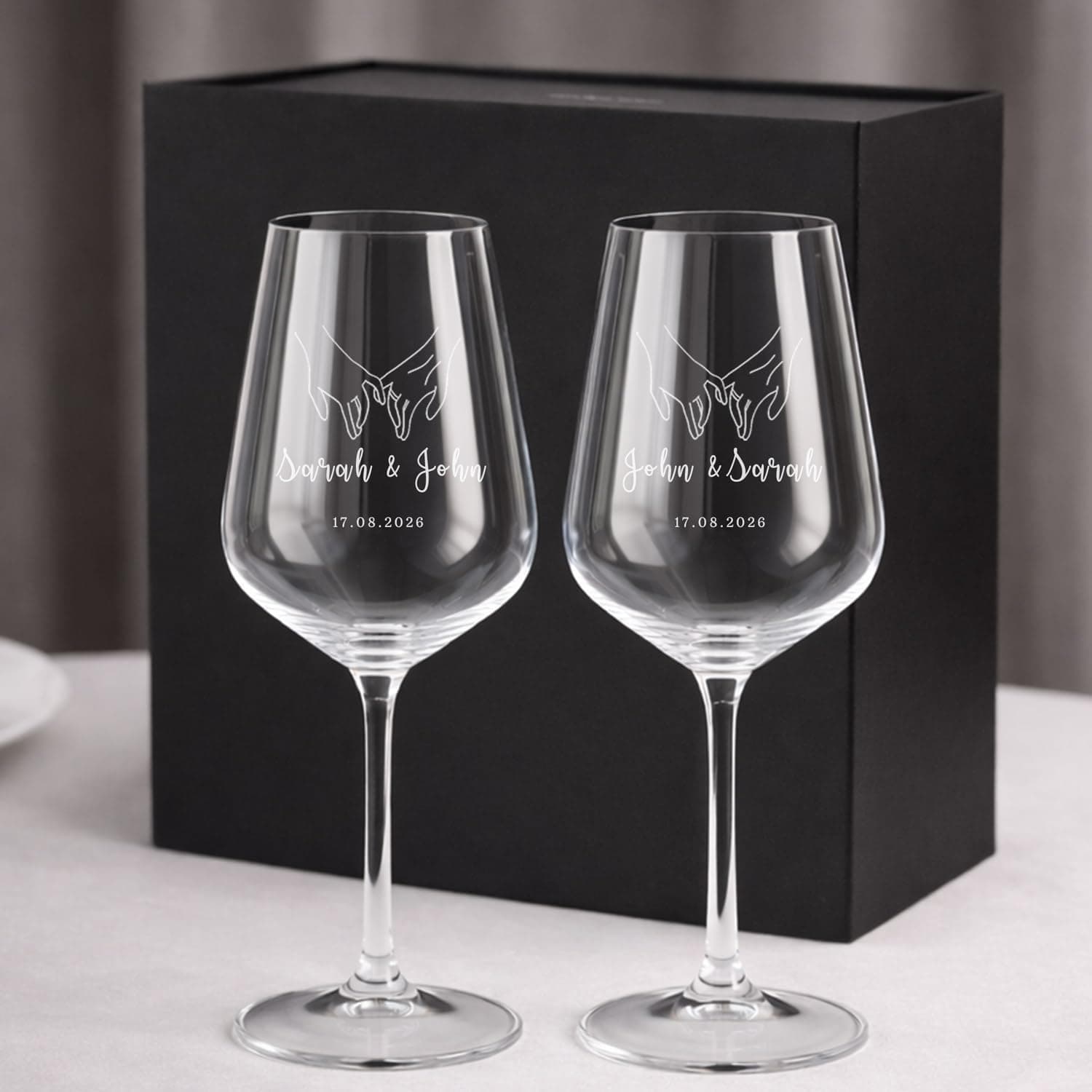 Dos copas de vino de cristal grabadas con dos manos entrelazadas y los nombres "Sarah & John" y "John & Sarah" con la fecha 17.08.2026, en una caja de regalo negra
