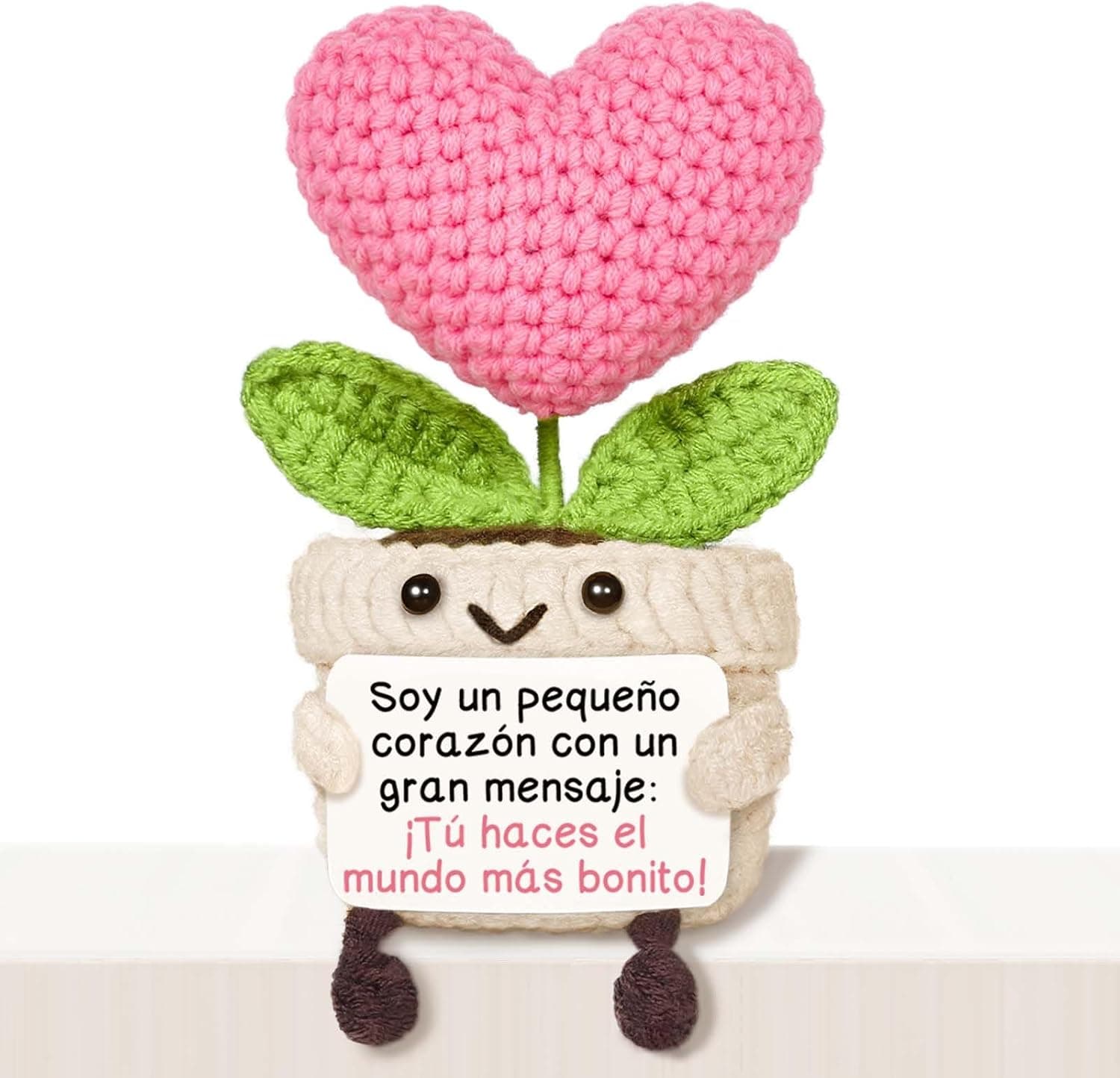 Muñeco de ganchillo con forma de corazón rosa sobre una maceta tejida con carita sonriente, sujetando un cartel con el mensaje