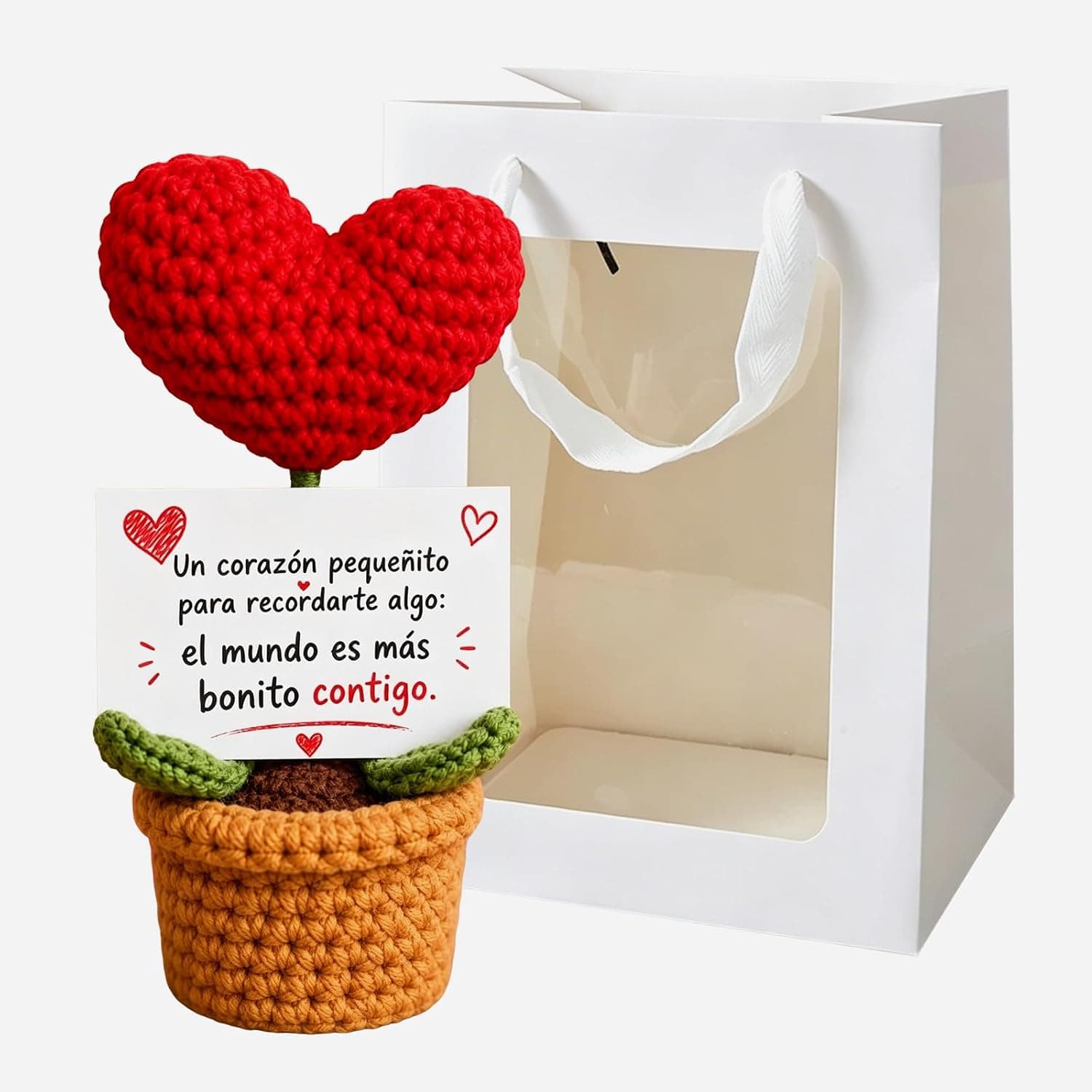 Corazón rojo de ganchillo en una macetita marrón con hojas verdes, junto a una tarjeta con el mensaje y una bolsa de regalo blanca