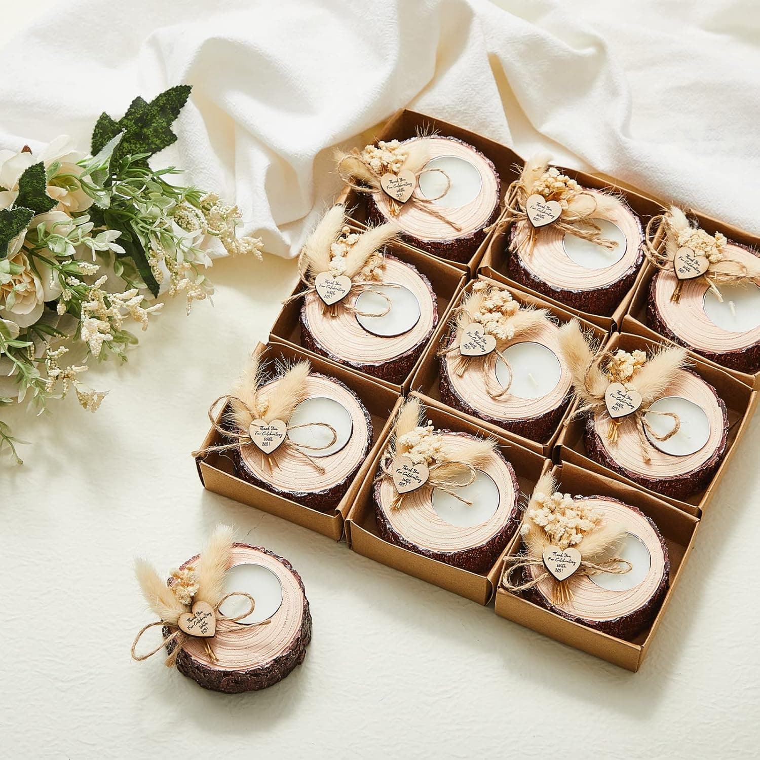 Nueve portavelas con forma de tronco de madera y vela de té encendida, cada uno decorado con flores secas, una etiqueta de agradecimiento y atado con cordel, colocados sobre tela blanca junto a un ramo de flores