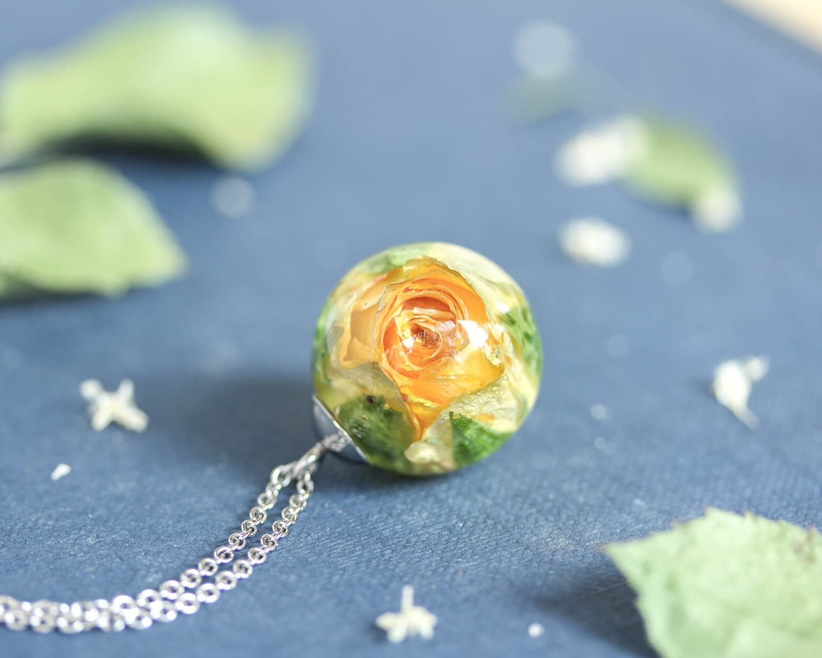 Handmade yellow rose resin pendant displayed on a dark blue background, surrounded by scattered dried flowers and leaves.